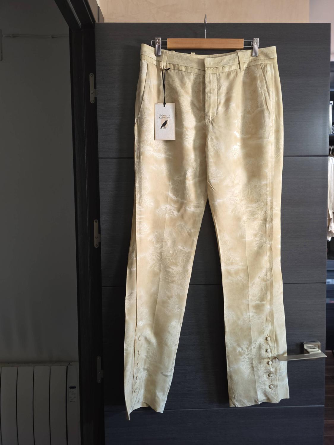 Light-coloured trousers with a subtle pattern and buttons at the hem hanging on a door. Label with the text “Halloween Collection” and a bird.