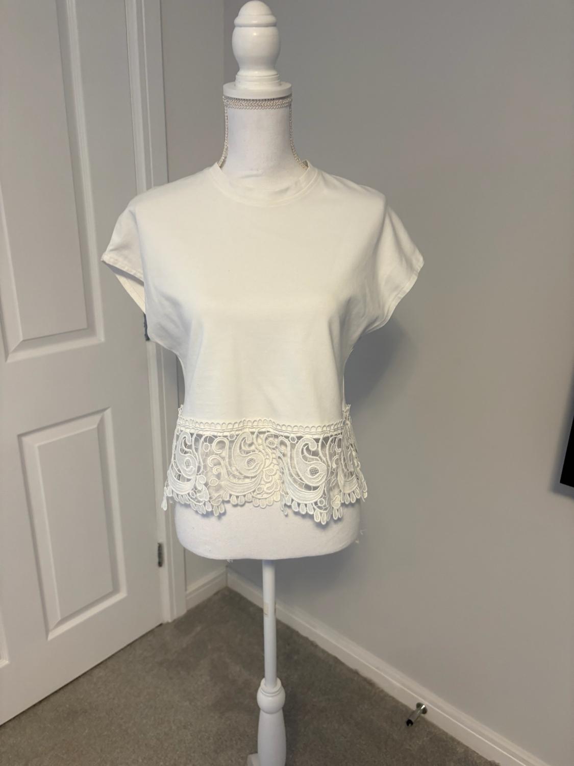 White short-sleeved T-shirt with a lace hem on a white mannequin, in a room with grey walls and a white door.