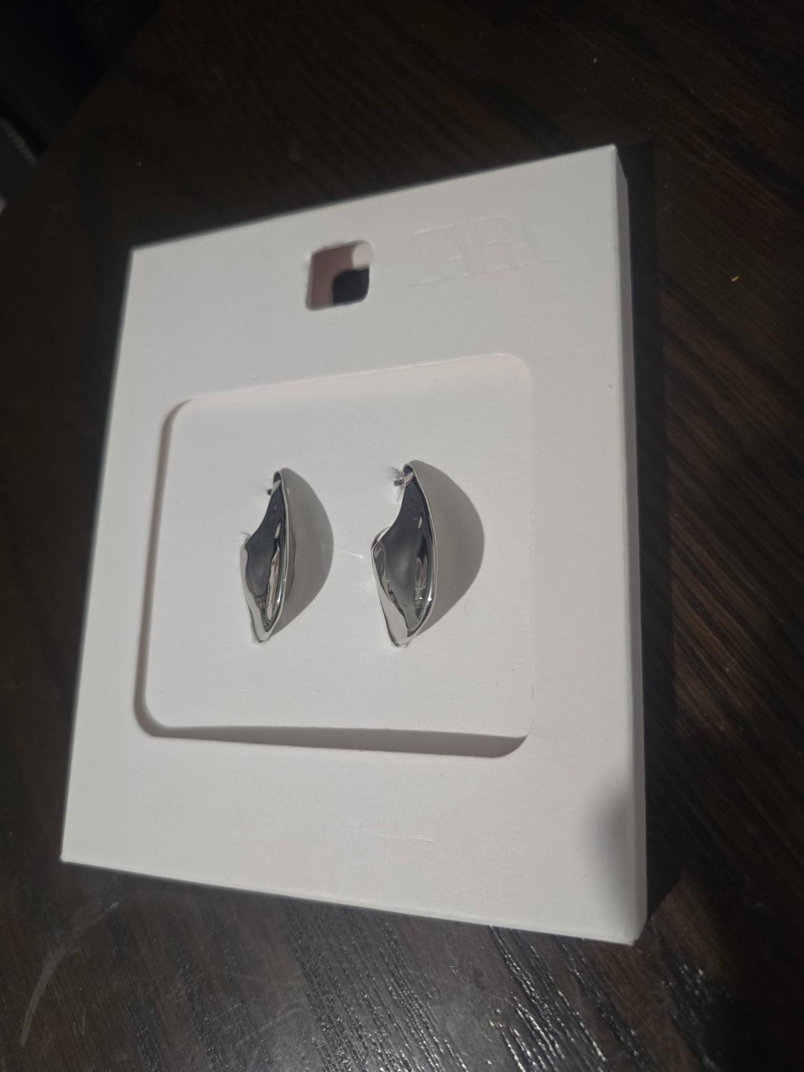 Pair of irregularly shaped silver-plated earrings on a white stand with the text ZARA, on a dark wooden surface.