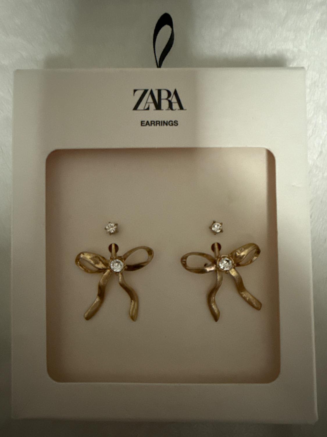 Gold bow-shaped earrings with rhinestone detail in a white box. Visible text: "ZARA EARRINGS".