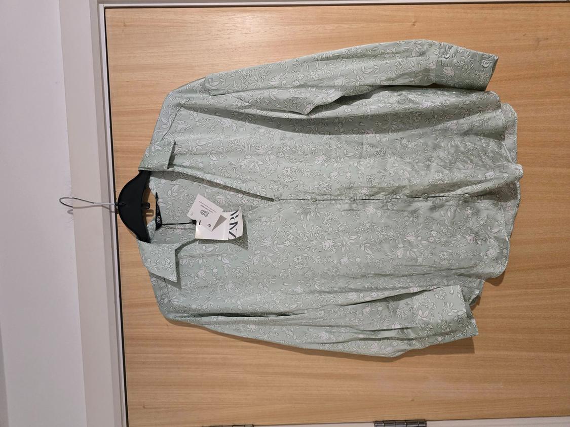 Light green shirt with floral print hanging on a black hanger over a wooden door. Labels visible on the hanger.