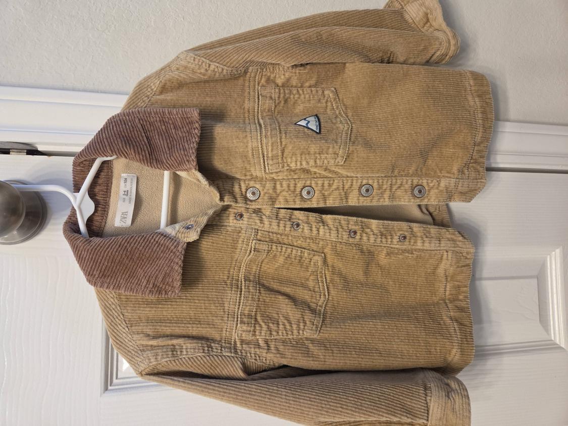 Beige corduroy jacket with brown collar hanging on a white hanger over a white door. Label visible on the inside.