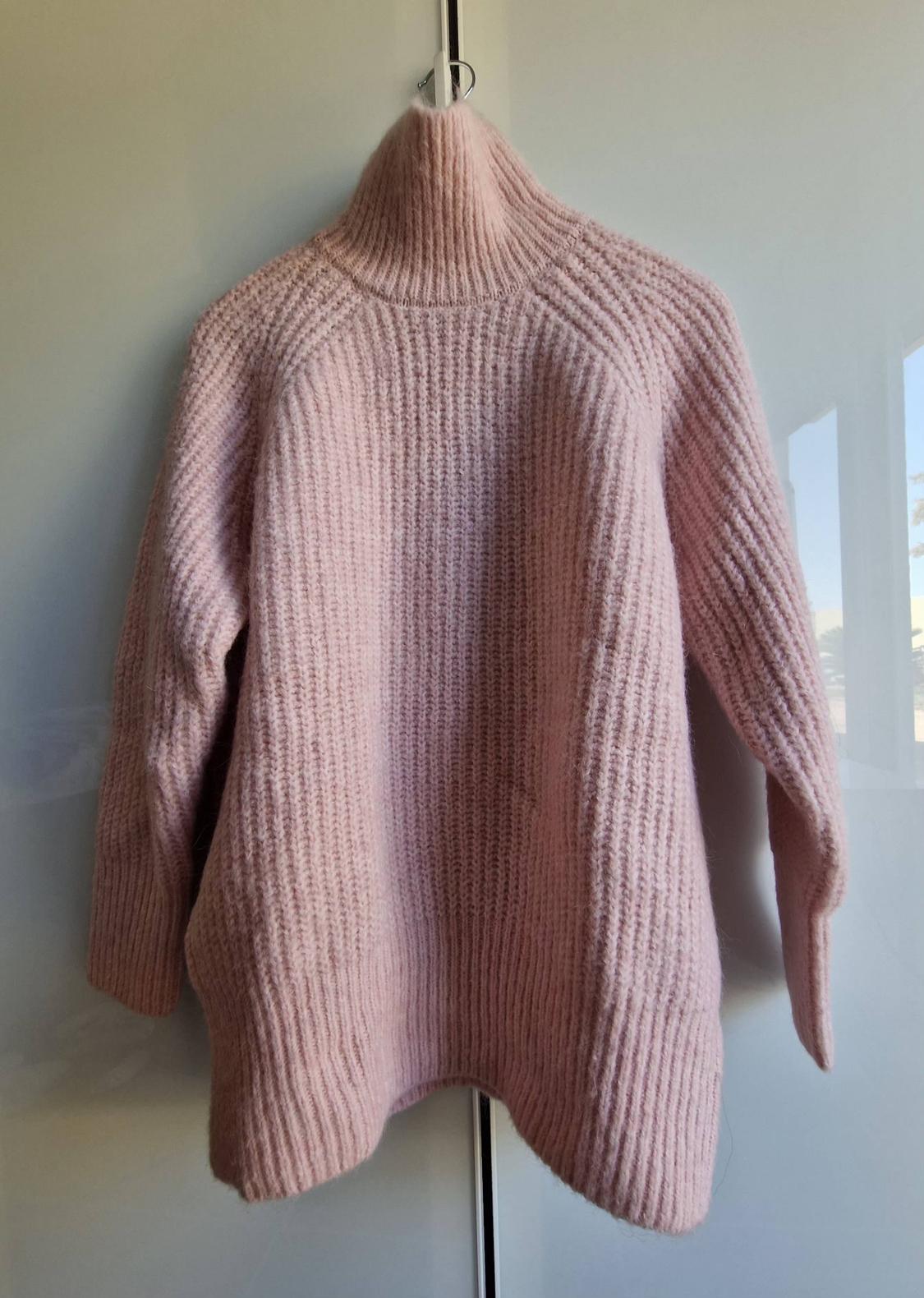 Light pink chunky-knit jumper with a high neck, hanging on a hanger against a white surface with natural light reflections.
