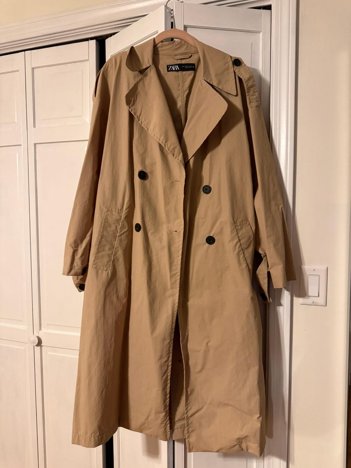 Zara beige trench coat hanging on a hanger in front of a white wardrobe. Classic design with black buttons and wide lapels.