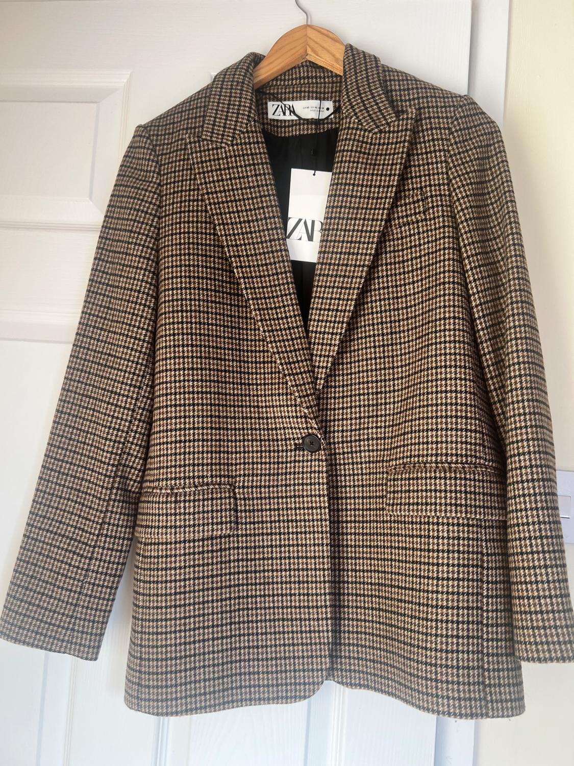 Brown and black checked jacket hanging on a wooden hanger. Visible label with the text "ZARA".