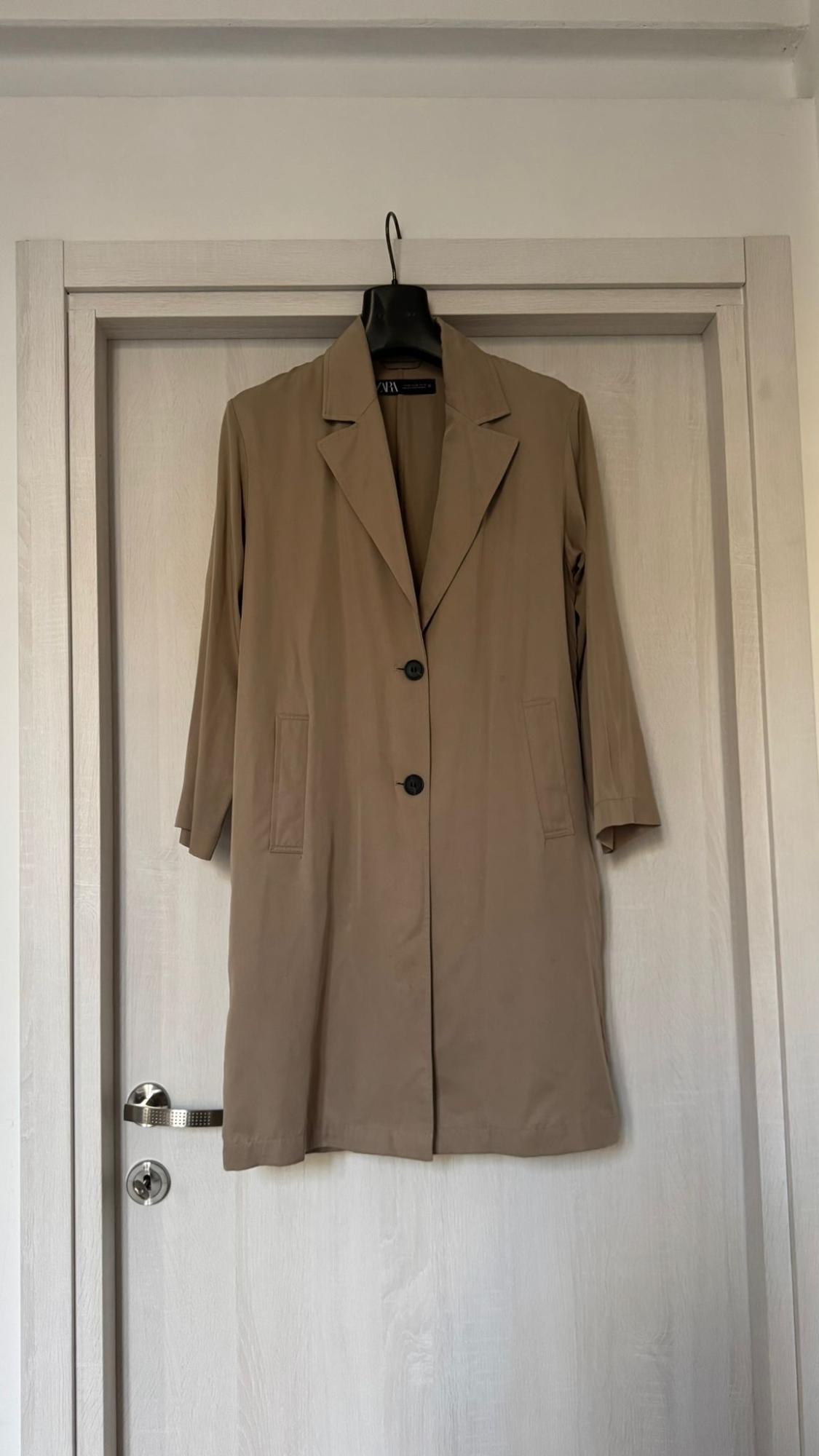 A beige coat hanging on a light wooden door. It has two black buttons and a lapelled collar.