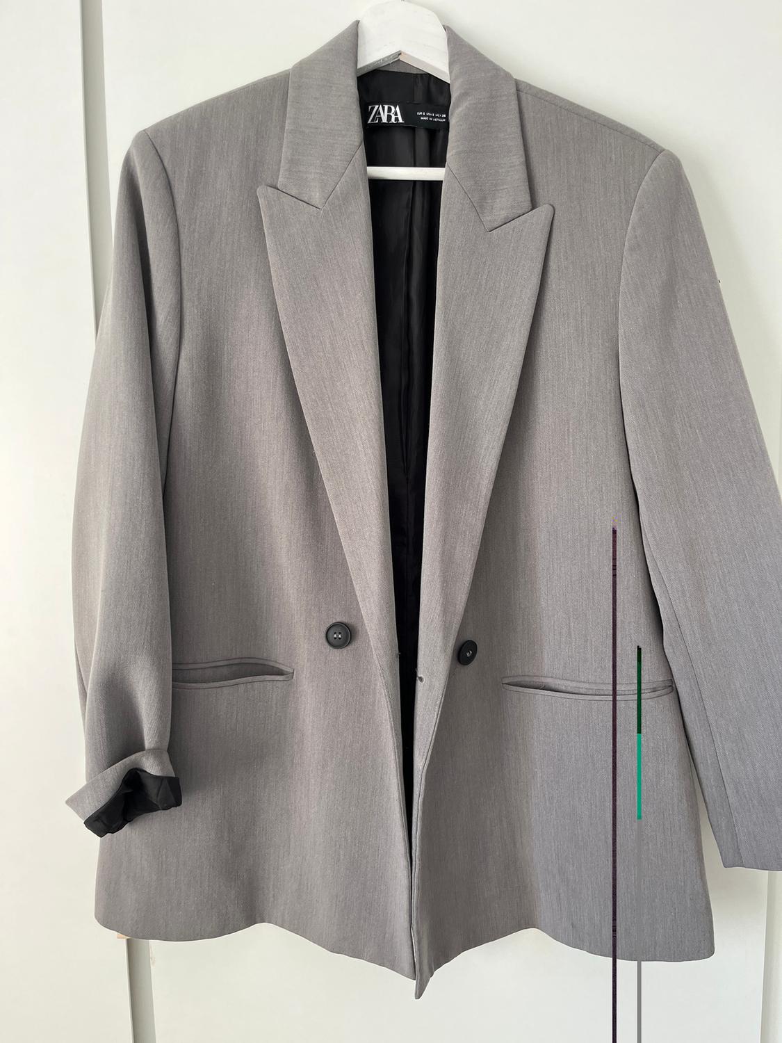 Grey suit jacket hanging on a white hanger. Visible tag with "ZARA" brand. White background.