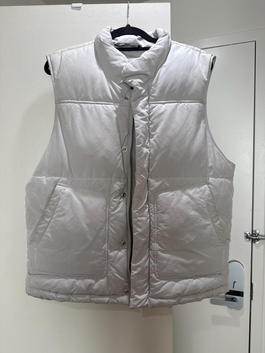 Light gray quilted vest with a high collar and front pockets, hanging on a black hanger in front of a white door.