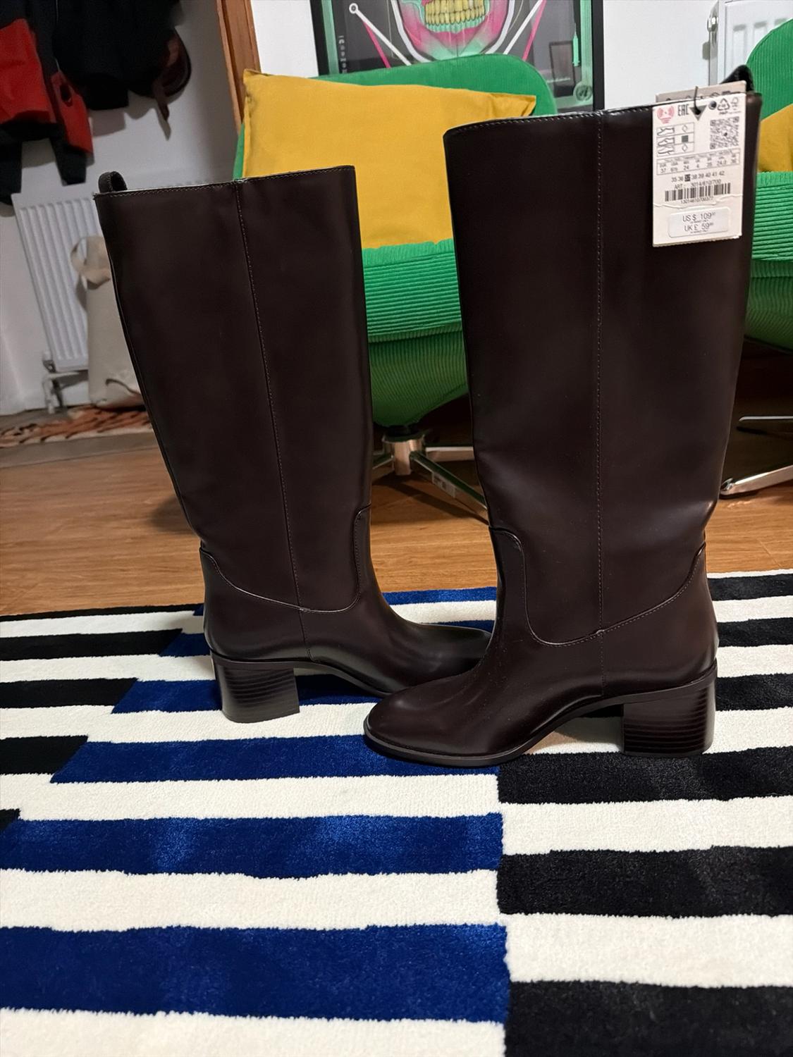 Tall brown heeled boots on a black, white and blue striped rug. Background with a green chair, yellow cushion and wooden floor.