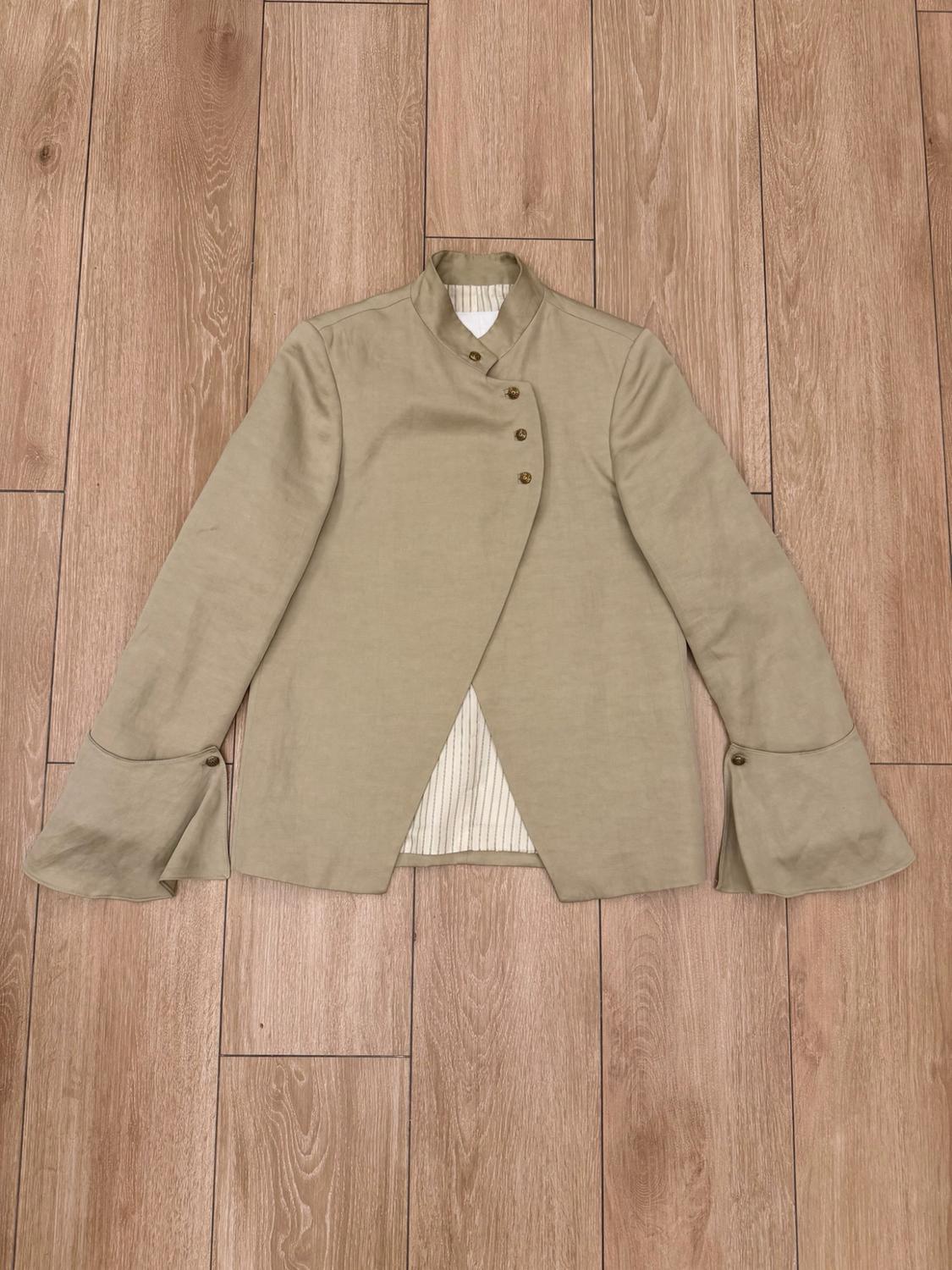 Beige high-neck jacket with an asymmetric fastening and flared sleeves with buttons, on a wooden floor. Striped lining.
