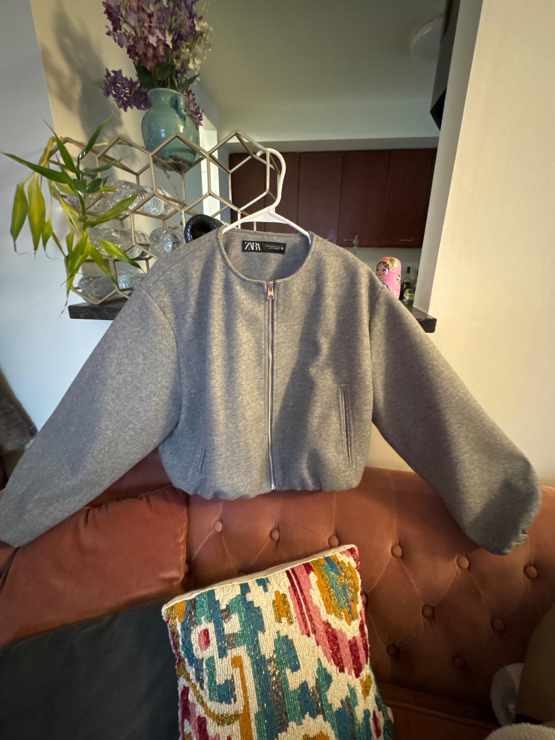 Gray zippered jacket hanging on a hanger over a pink sofa. In the background, a shelf with plants and a blue vase.