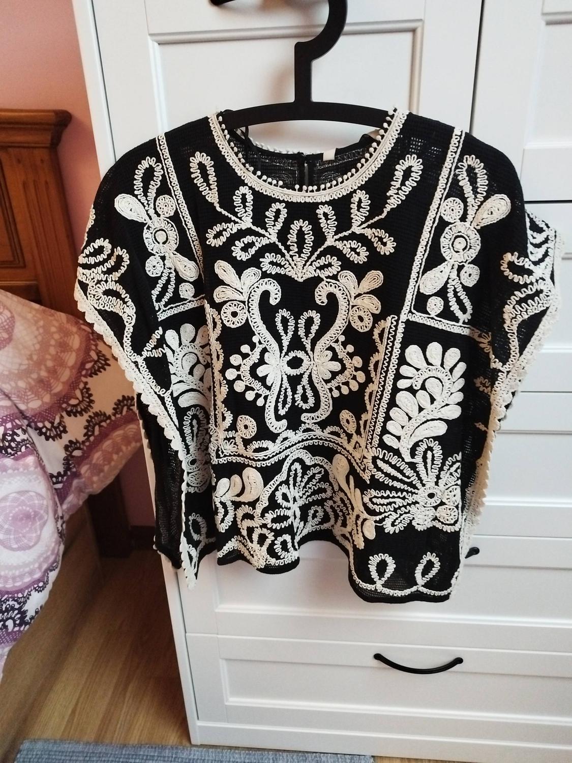 Black blouse with white embroidery hanging on a black hanger in front of a white wardrobe. Background with bed and pink wall.