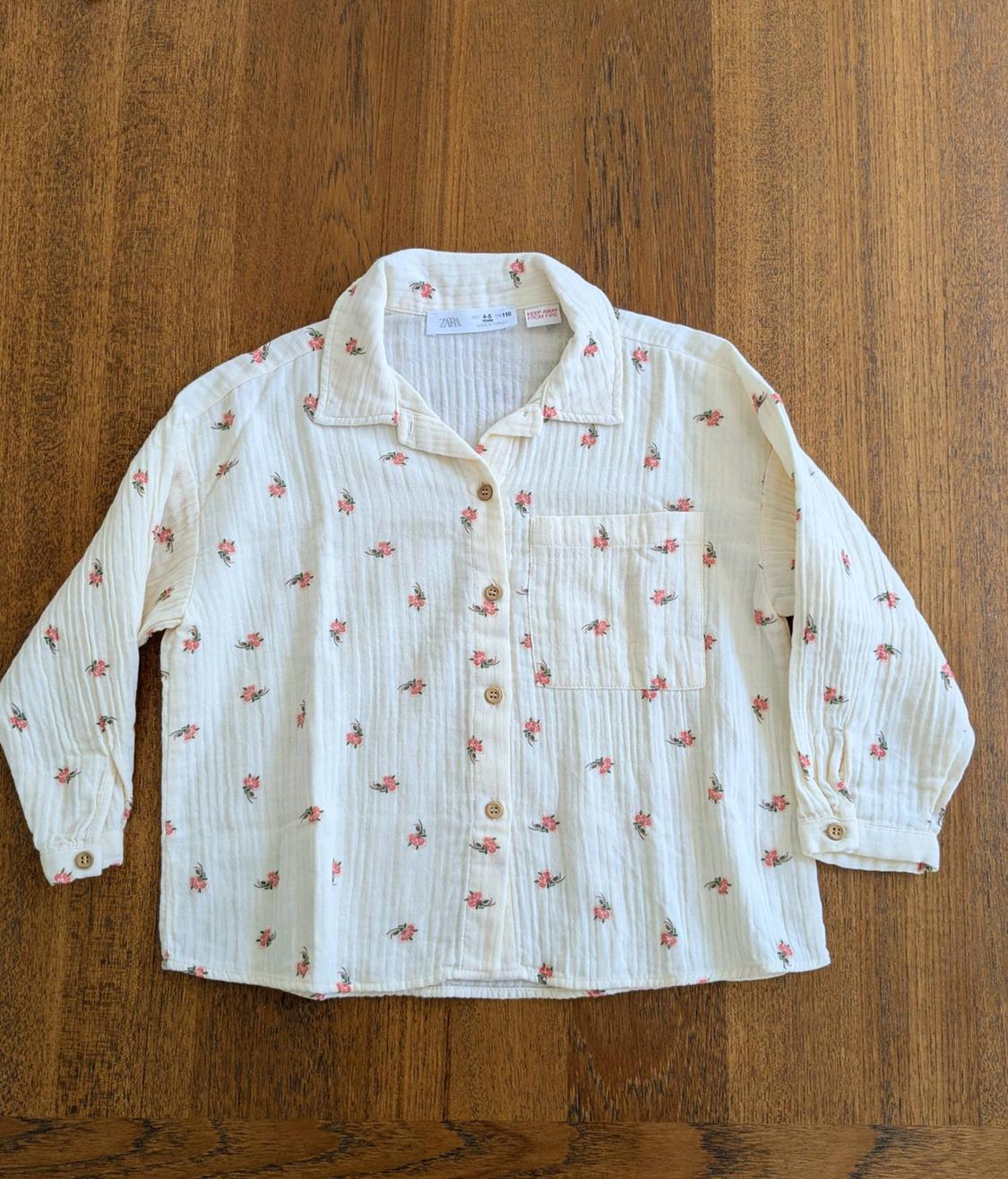 White long-sleeved shirt with a small red floral print and wooden buttons, on a wooden surface.