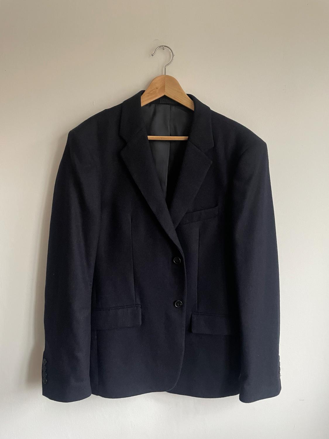 Black jacket hanging on a wooden hanger against a white wall. Classic design with lapels and two front buttons.