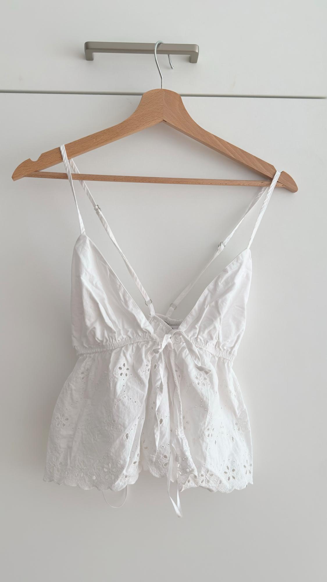 White camisole top with embroidered details, hanging on a wooden hanger against a white background.