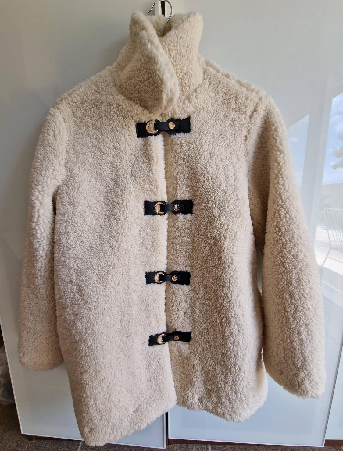 Cream-coloured textured curly coat hanging up. It has a high neck and four black front fastenings with gold-coloured metal rings.