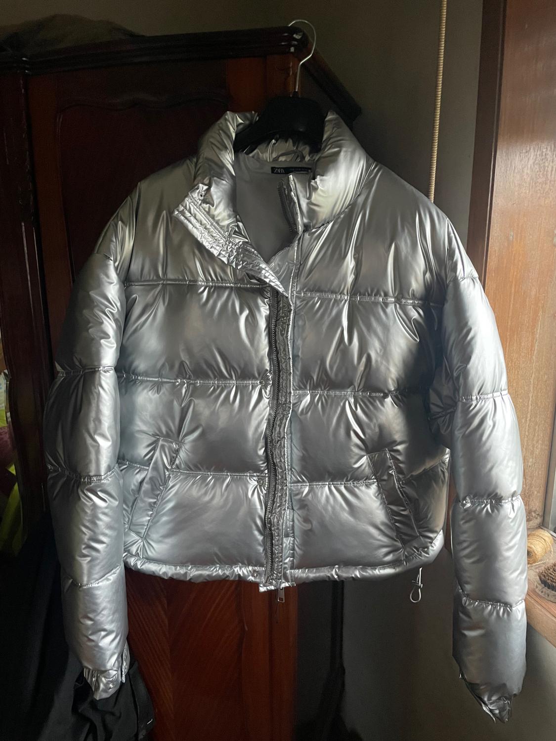 Silver padded jacket hanging on a hanger, with a front zip and pockets, on a dark wooden background.