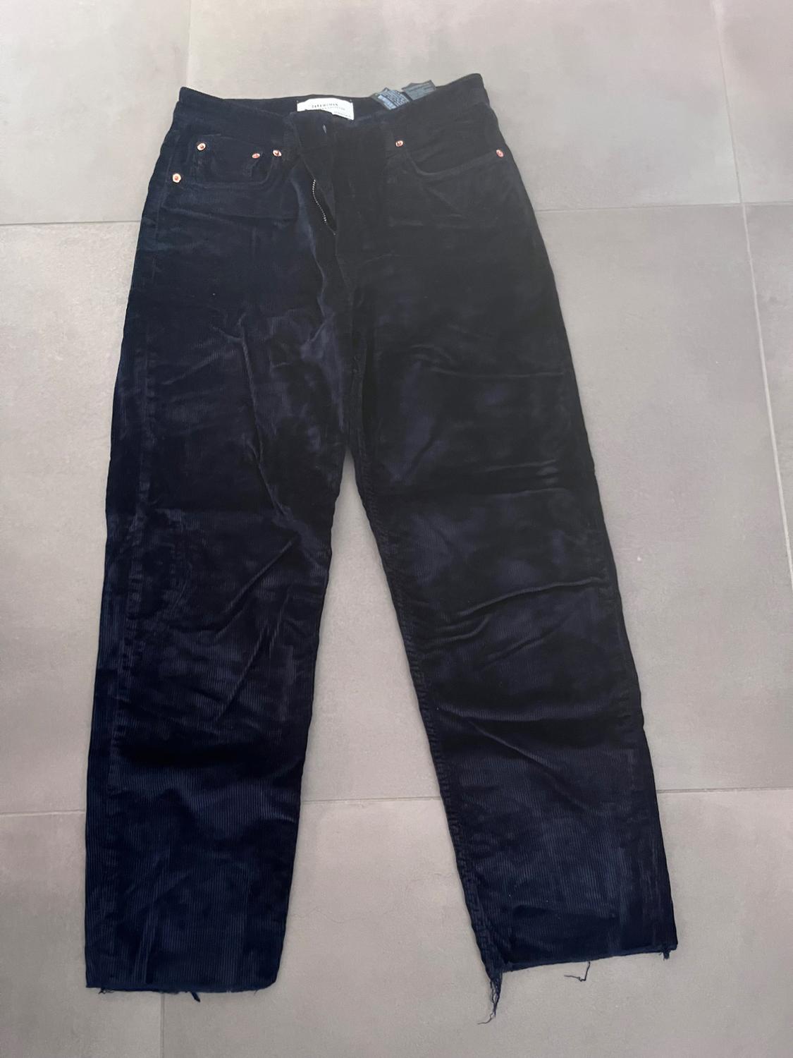 Black corduroy trousers with front pockets and metallic details, placed on a light grey background.
