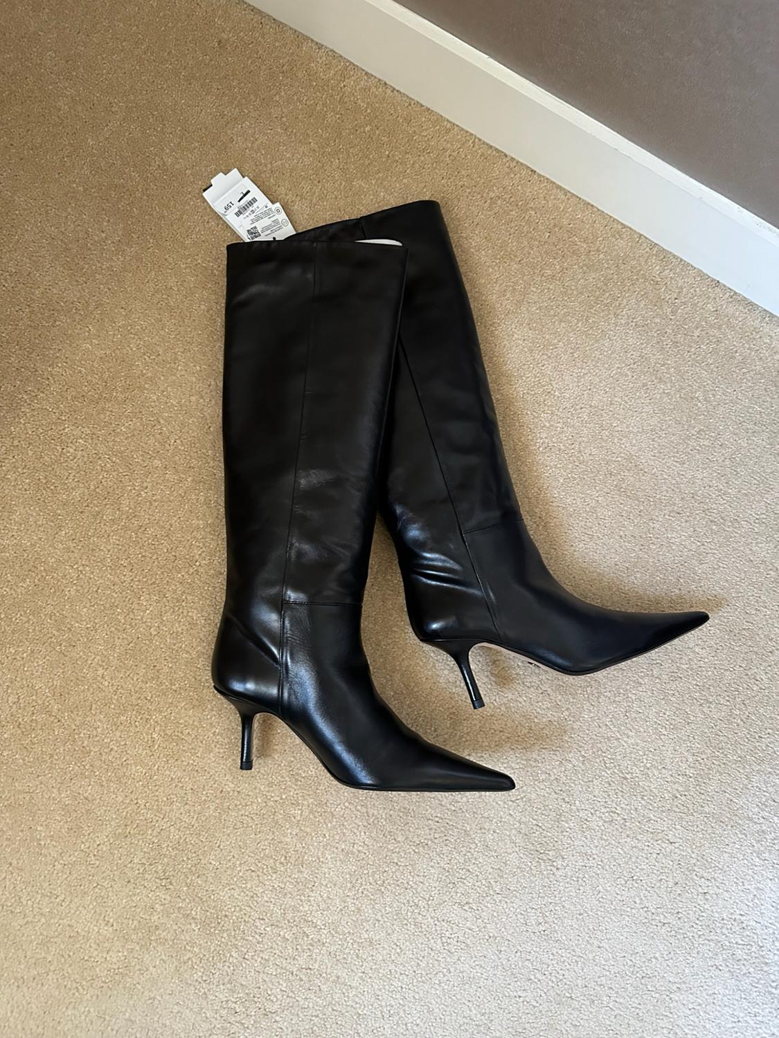 Black knee-high boots with slim heels and pointed toes on a beige carpet beside a white skirting board. They have a label hanging from them.