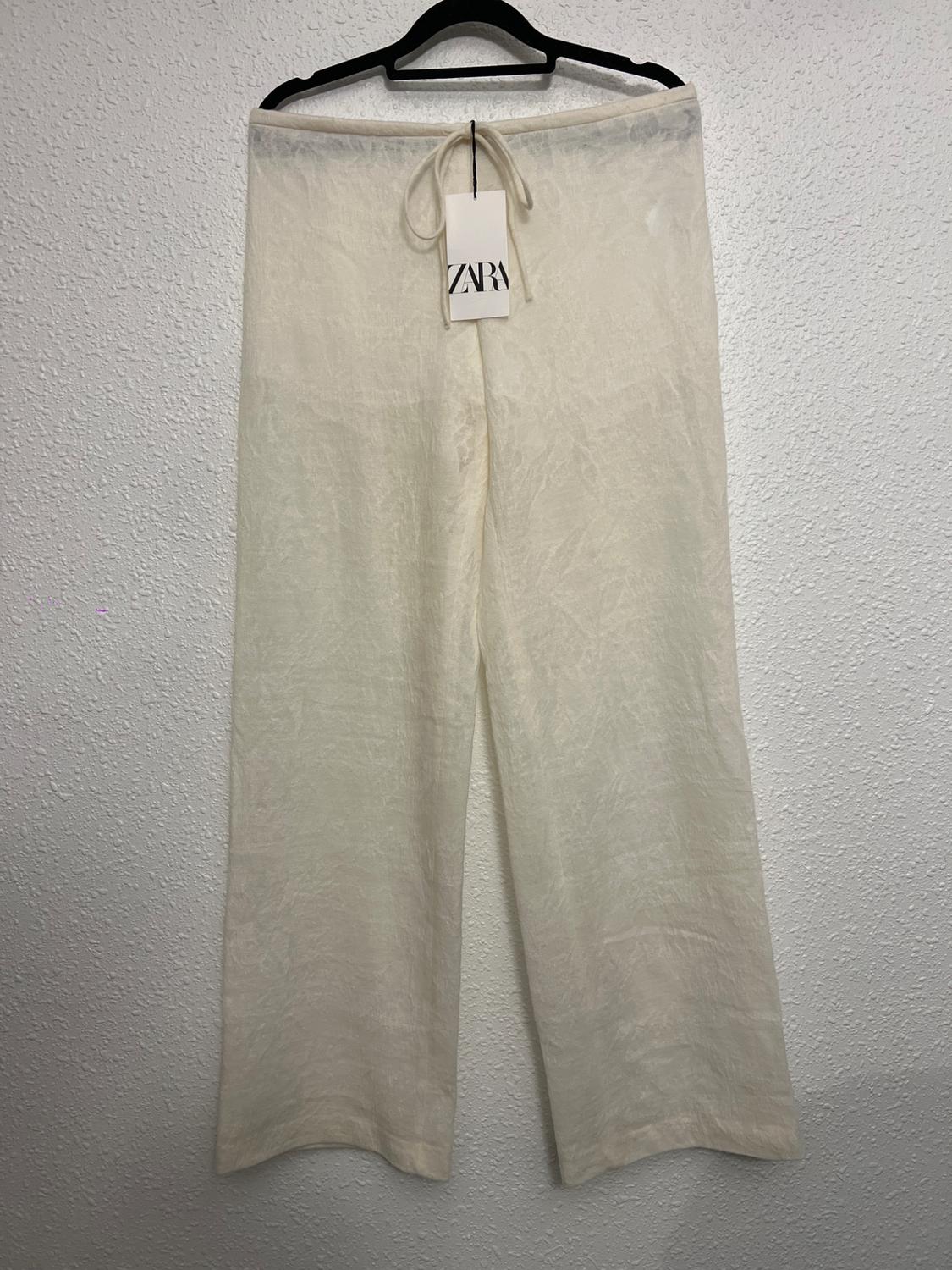 White trousers hanging on a black hanger with a Zara tag. Textured white wall background.