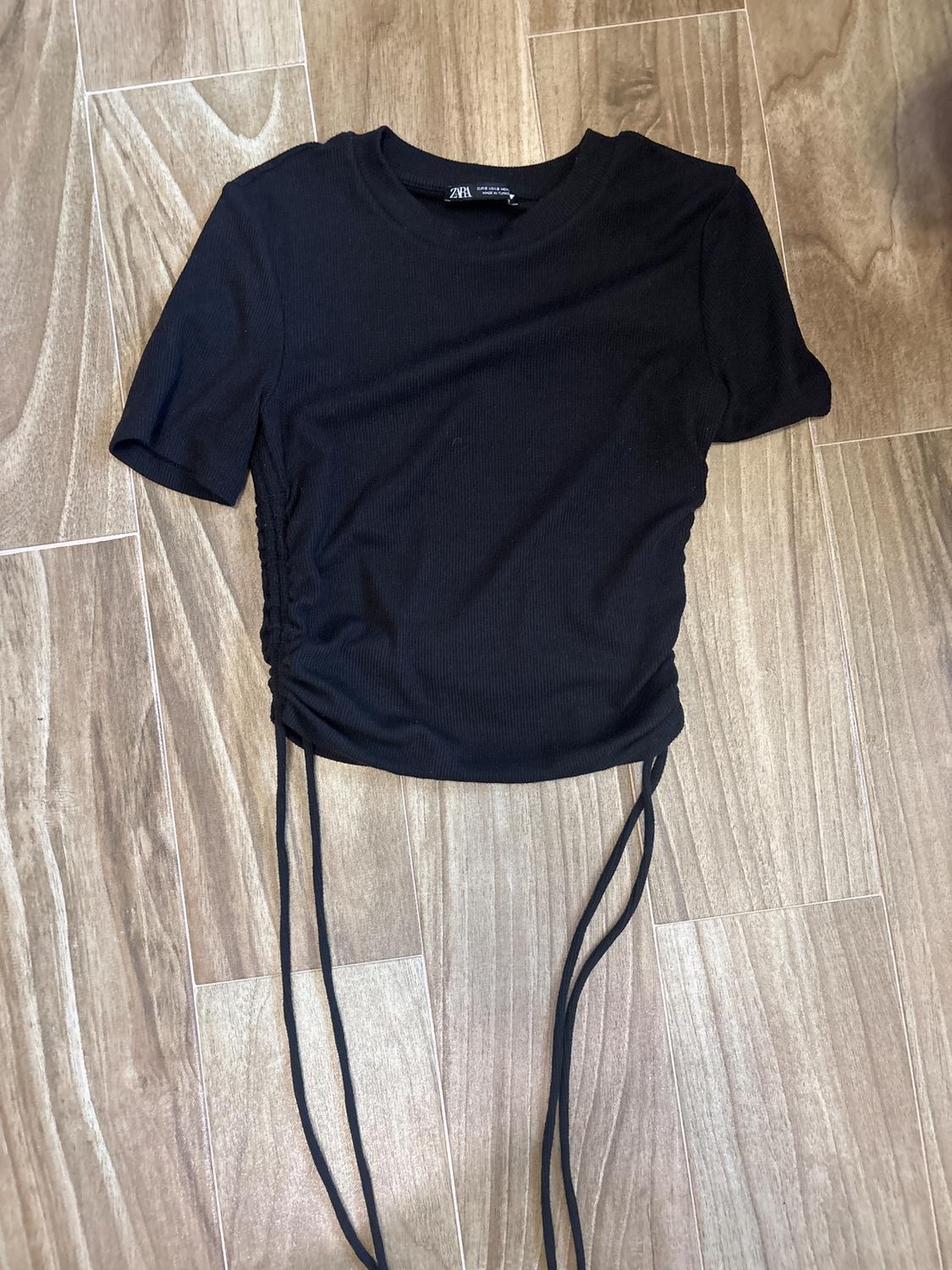 Black short-sleeved t-shirt with adjustable laces on the sides, on a light wooden floor. Visible label on the collar.