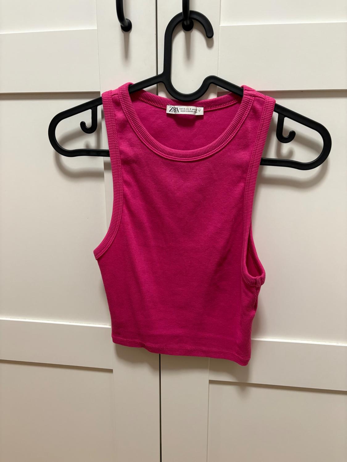 Pink ribbed sleeveless crop top hanging on a black hanger on a white door. The label reads ZARA.