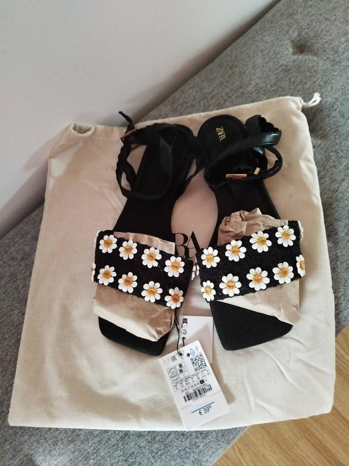 Black sandals with white and yellow flowers on a beige bag. Visible tag with price of €39.95.
