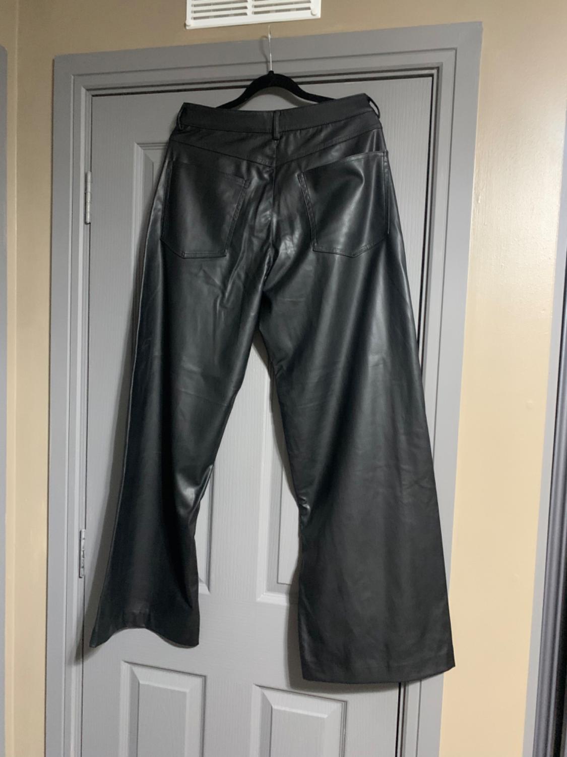 Black leather trousers hanging on a hanger over a grey door. The image shows the back of the trousers.