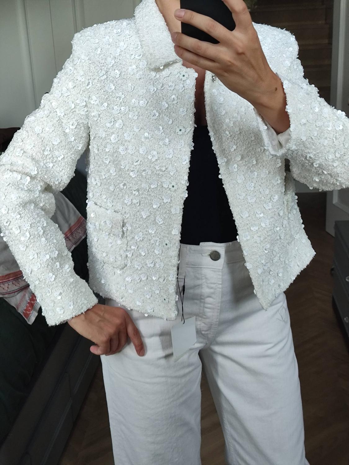 Mirror selfie of a person in a white sequinned jacket, black top and white trousers with a visible tag in a room.
