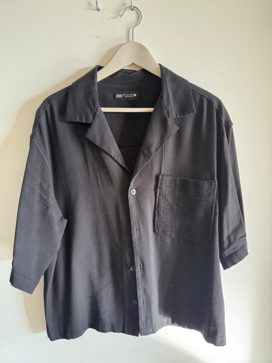 Black short-sleeved shirt hanging on a wooden hanger. It has a textured pocket on the left side of the chest.