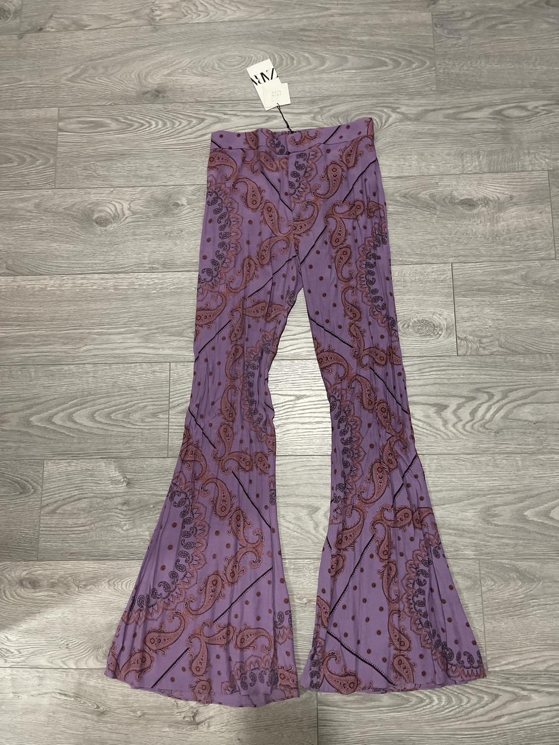 Purple flared trousers with paisley print and brown polka dots on a grey wooden floor. Visible label at the waist.