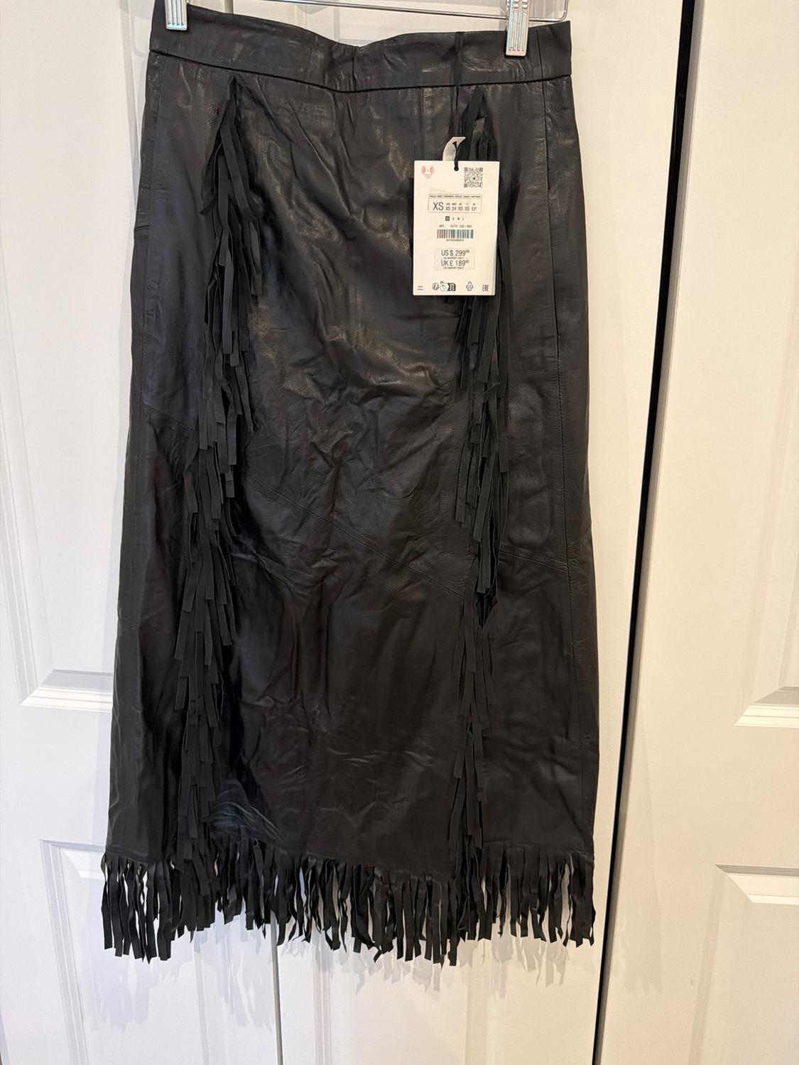 Black fringe skirt hanging on a hanger in front of a white door. Text: XS US $ 299 UK £ 189.