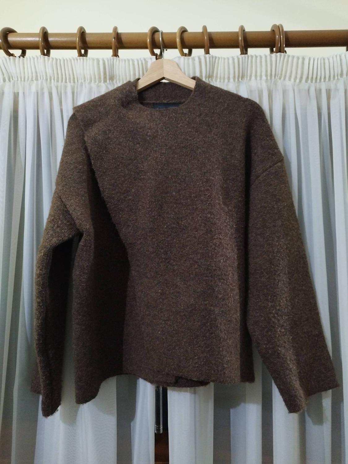 Long-sleeved brown jumper hanging on a wooden hanger in front of white curtains.