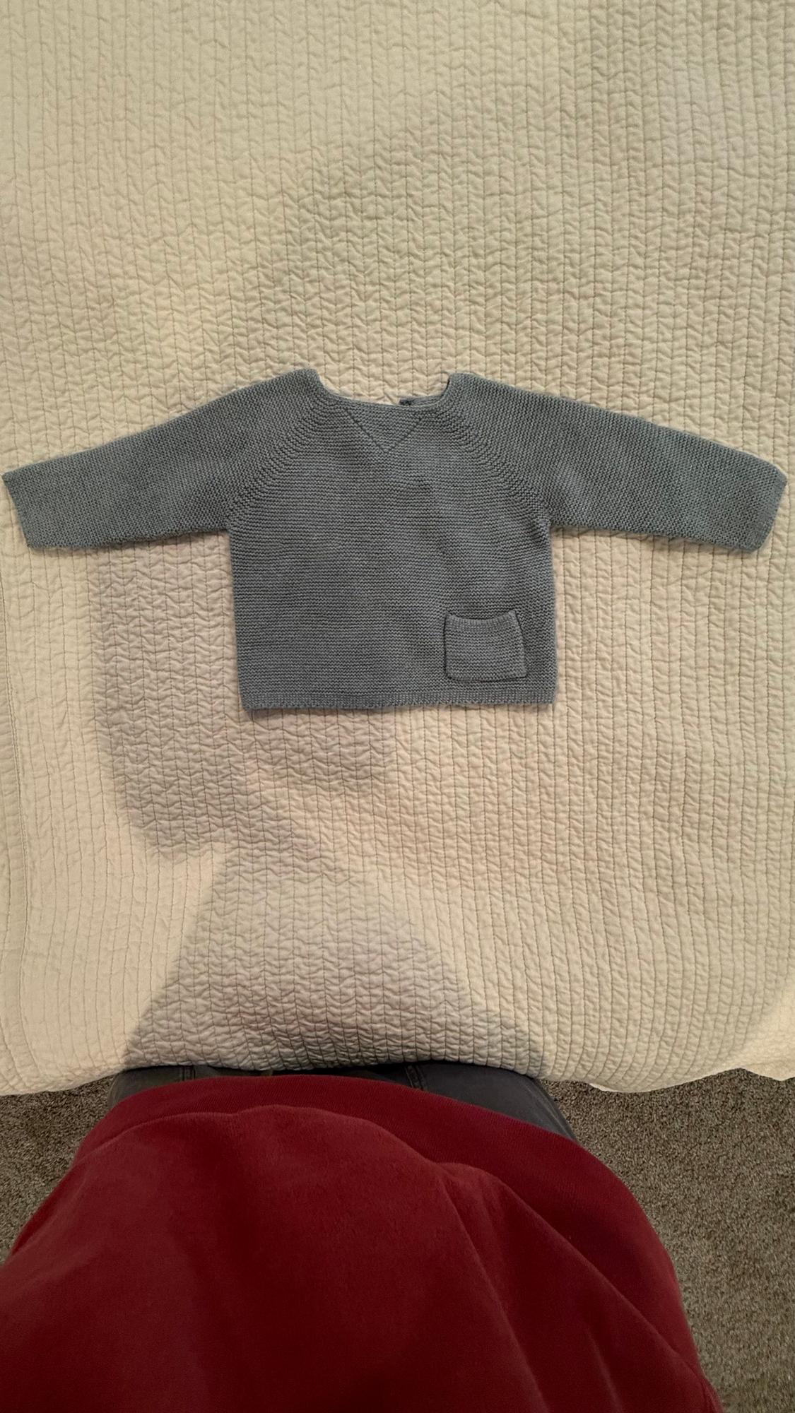 Grey knitted baby jumper with a small pocket, laid out on a textured beige quilt. Bottom of a red garment visible.