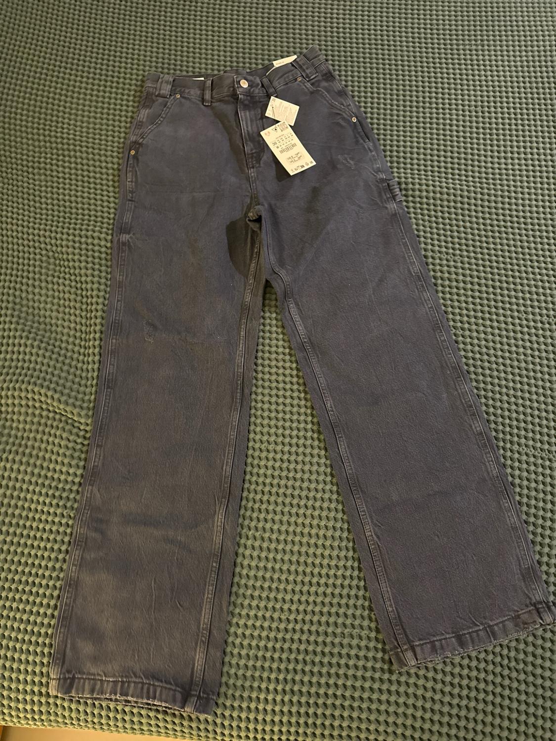 Dark blue wide-leg jeans with tags, laid out on a textured green surface. Casual and relaxed style.