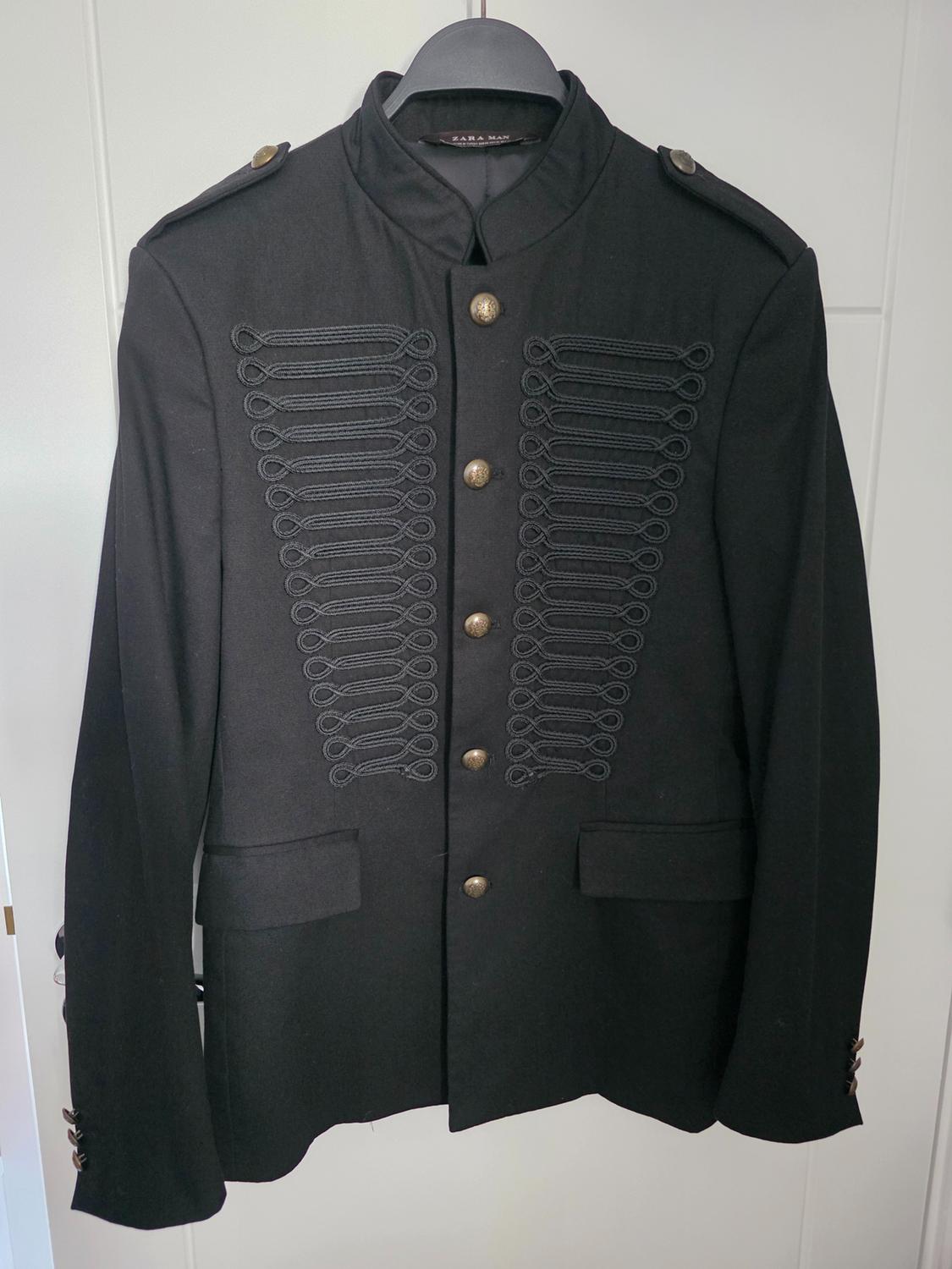 Black military-style jacket with gold buttons and lace-up details on the front, hanging on a black hanger.