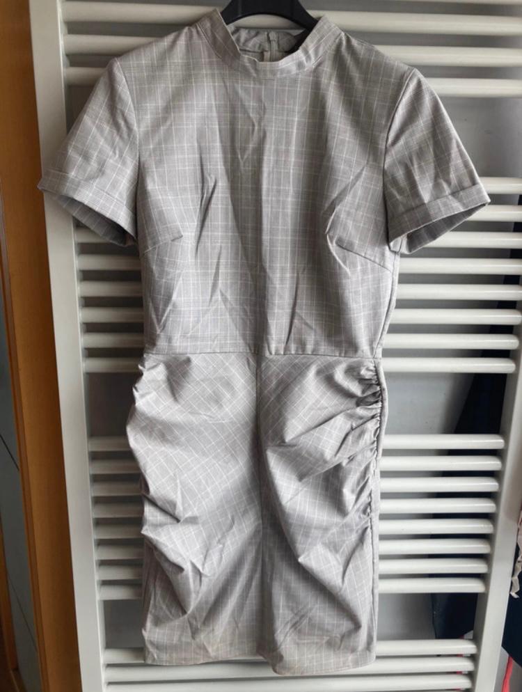 Grey short-sleeved dress with a check print, hanging on a black hanger against a background of white slats.