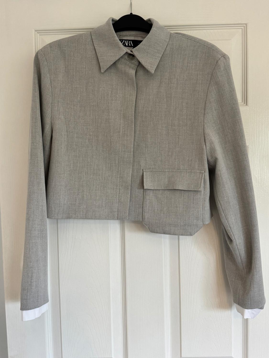 Short grey jacket with asymmetrical front pocket, classic collar and long sleeves, hanging on a hanger in front of a white door.