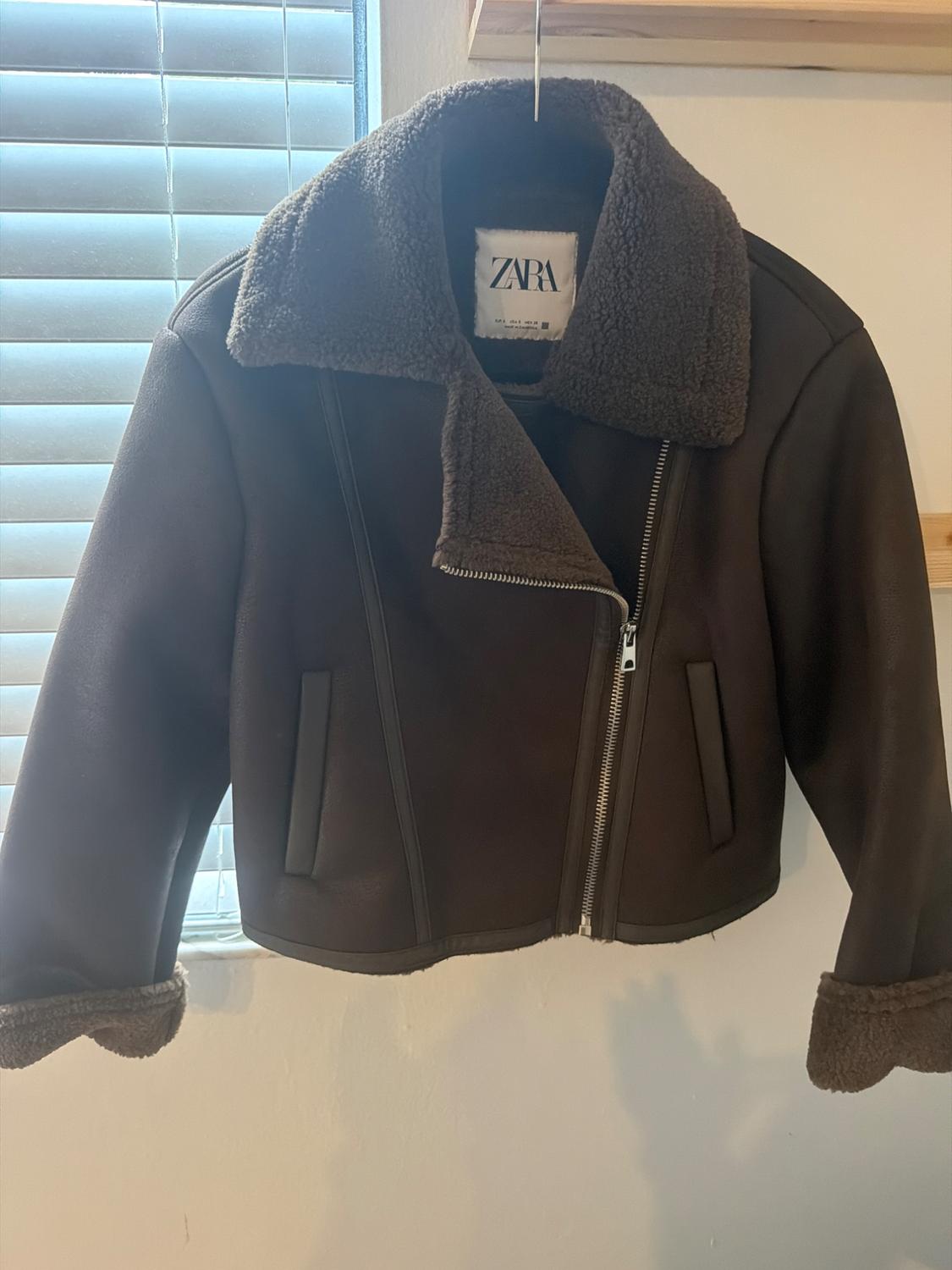 Dark brown jacket with a fur collar and asymmetrical zipper hanging on a hanger next to a blind. Label with the text ZARA.