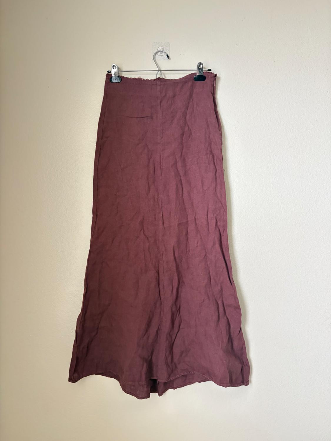 Burgundy long skirt in crinkled fabric with a frayed top edge. It is hanging from a clip hanger on a white wall.