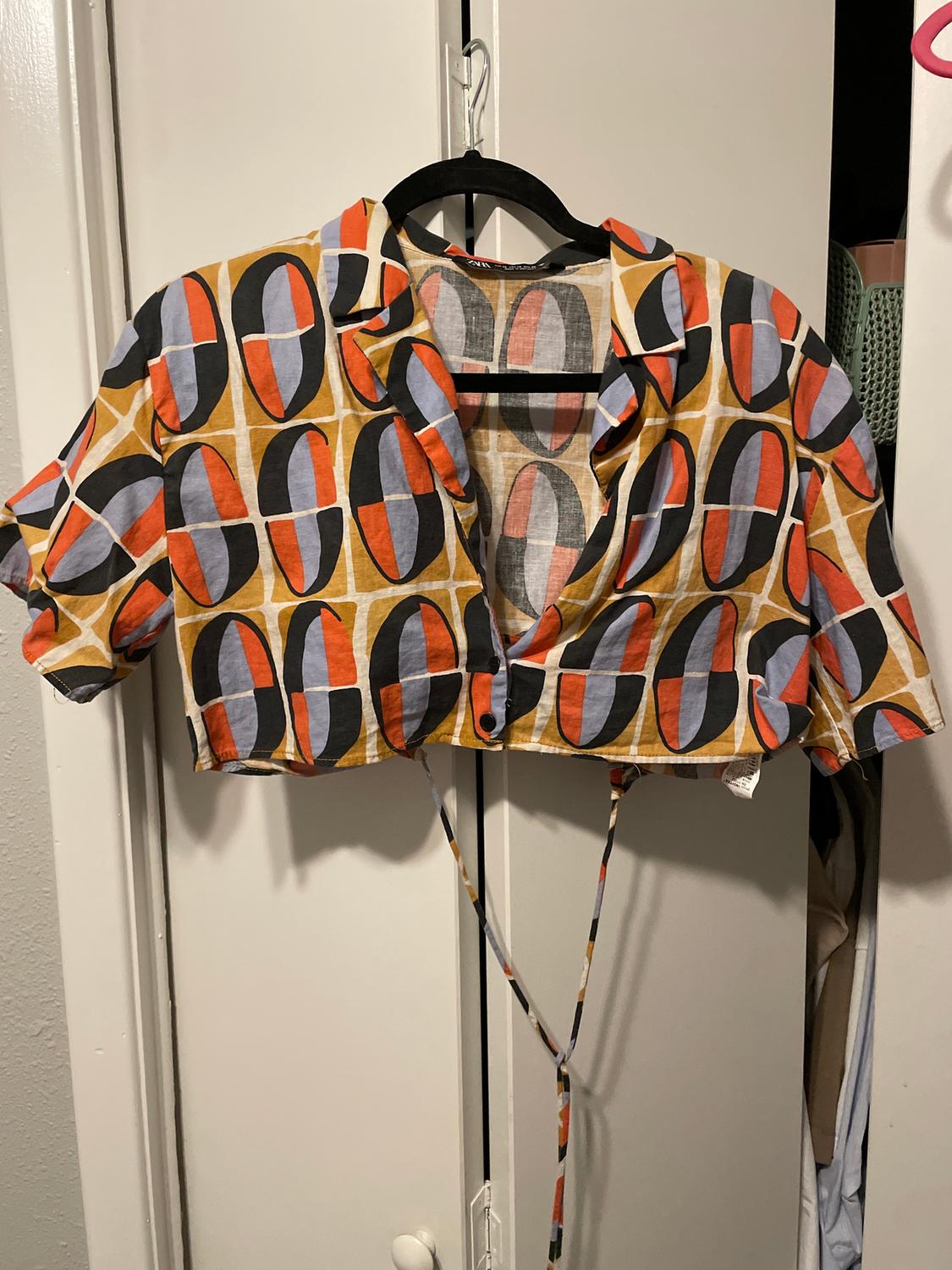 Cropped blouse with a geometric print in orange, black, and beige tones, hanging on a hanger in front of a white wardrobe.