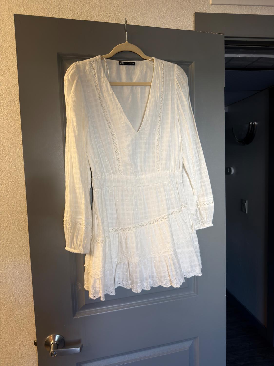 Short white dress with embroidery, long sleeves, V-neck, and ruffles. Hanging on a grey door. Casual style. Text: ZARA.