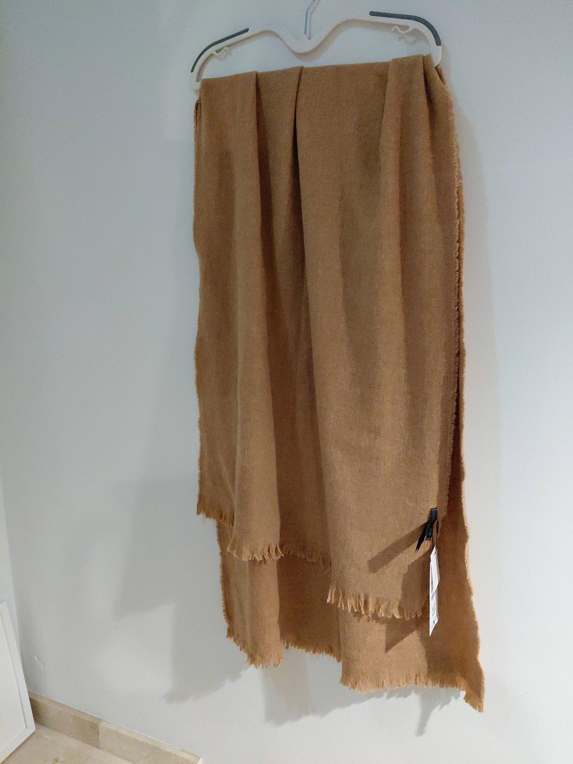 Long camel-coloured scarf with a fringed finish, hanging on a white coat hanger against a plain wall.