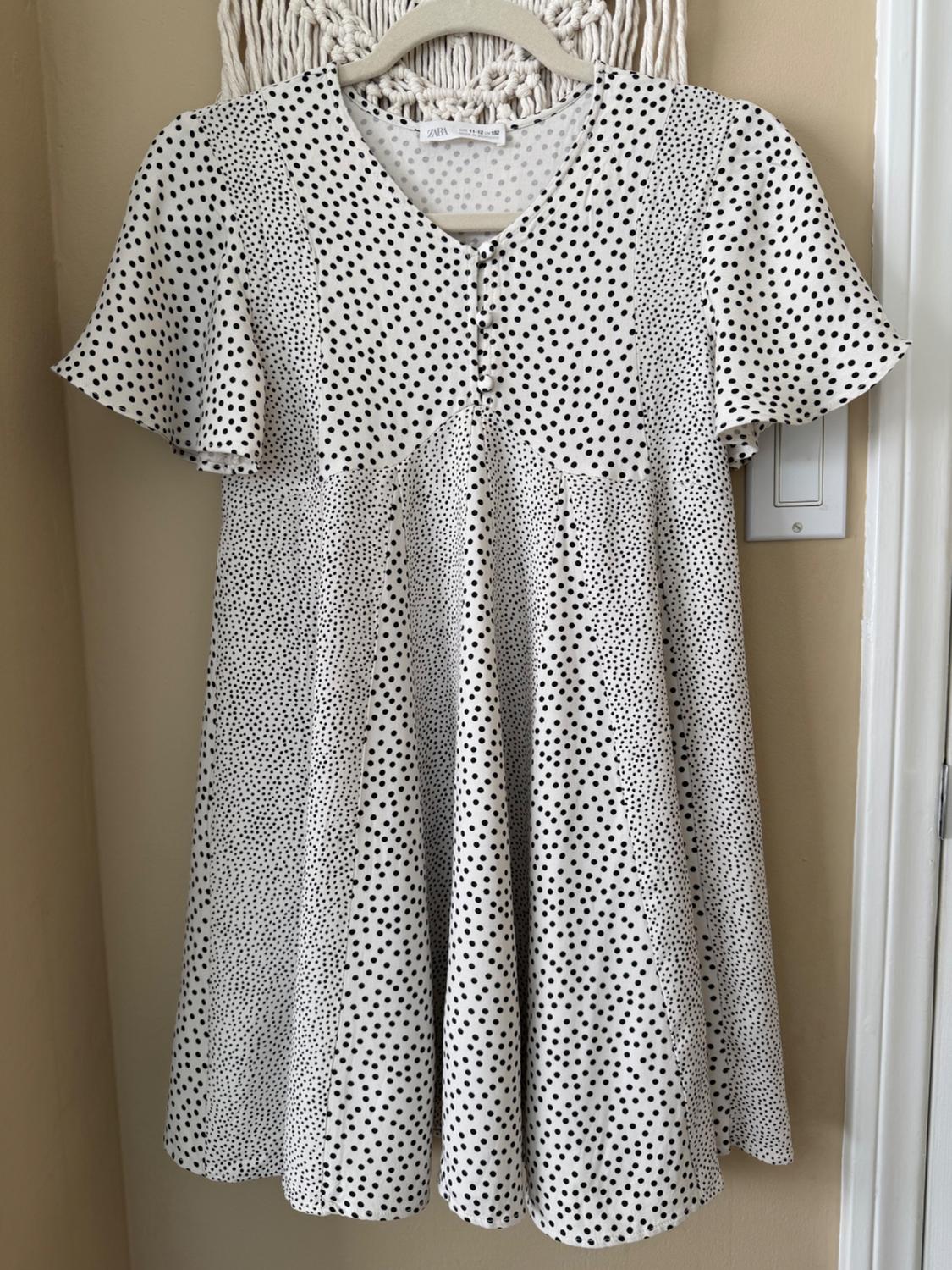 White dress with black polka dots hanging on a hanger. Short sleeves and V-neck. Beige background and wall with visible light switch.