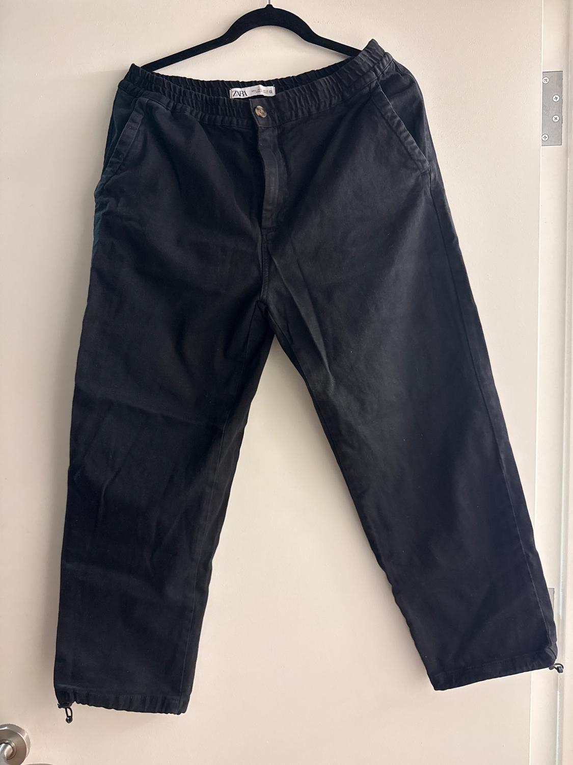 Black trousers hanging on a hanger over a white door. They have an elasticated waist and button fastening. Label visible on the inside.
