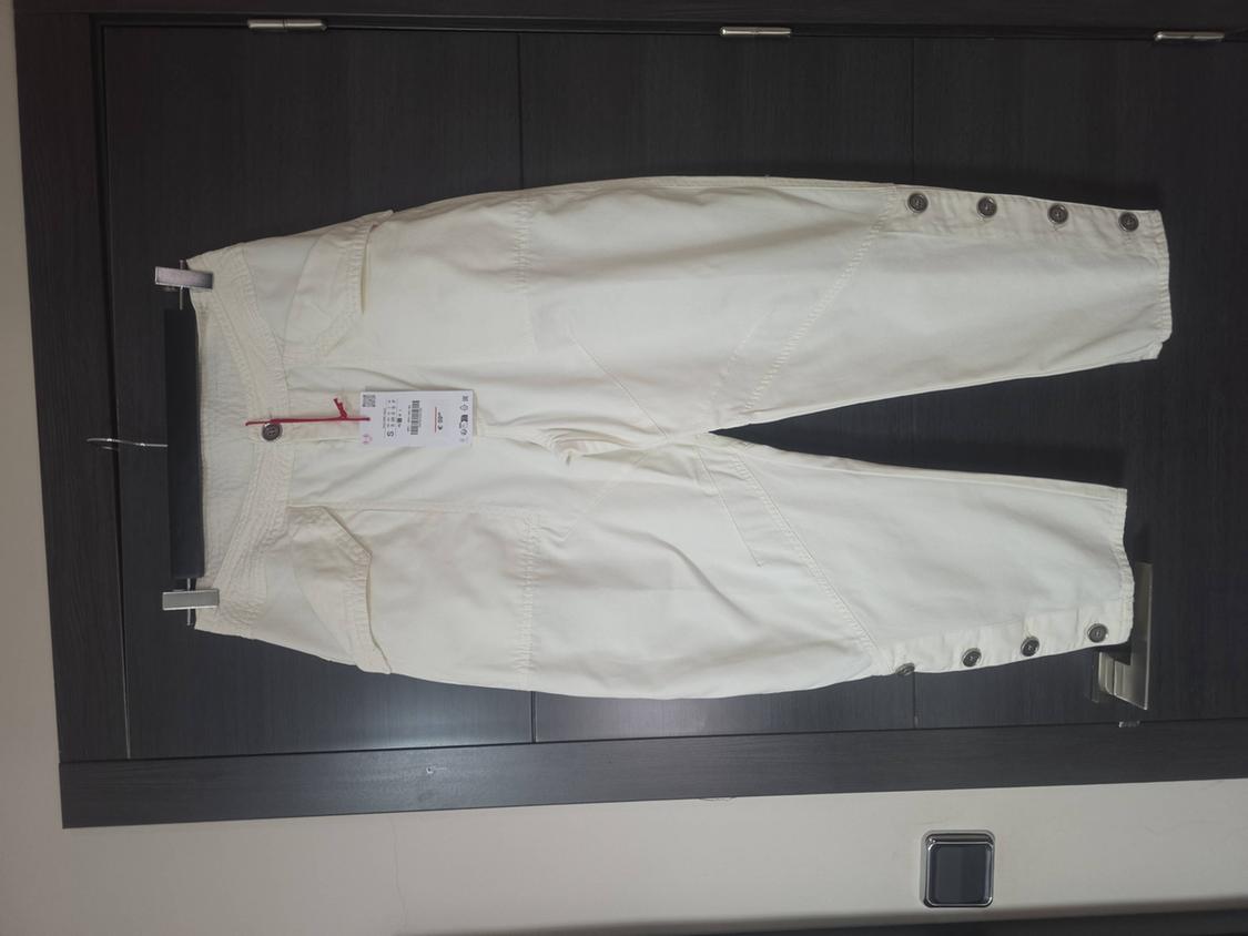 White capri trousers with buttons at the hem, hanging on a hanger on a dark door. Casual style with the label still attached.