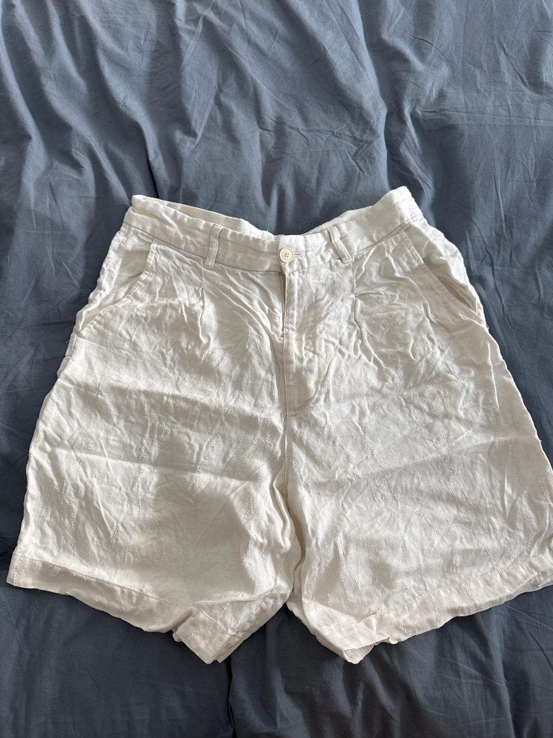 White crinkled shorts on a bed with grey sheets. White button visible on the waistband.