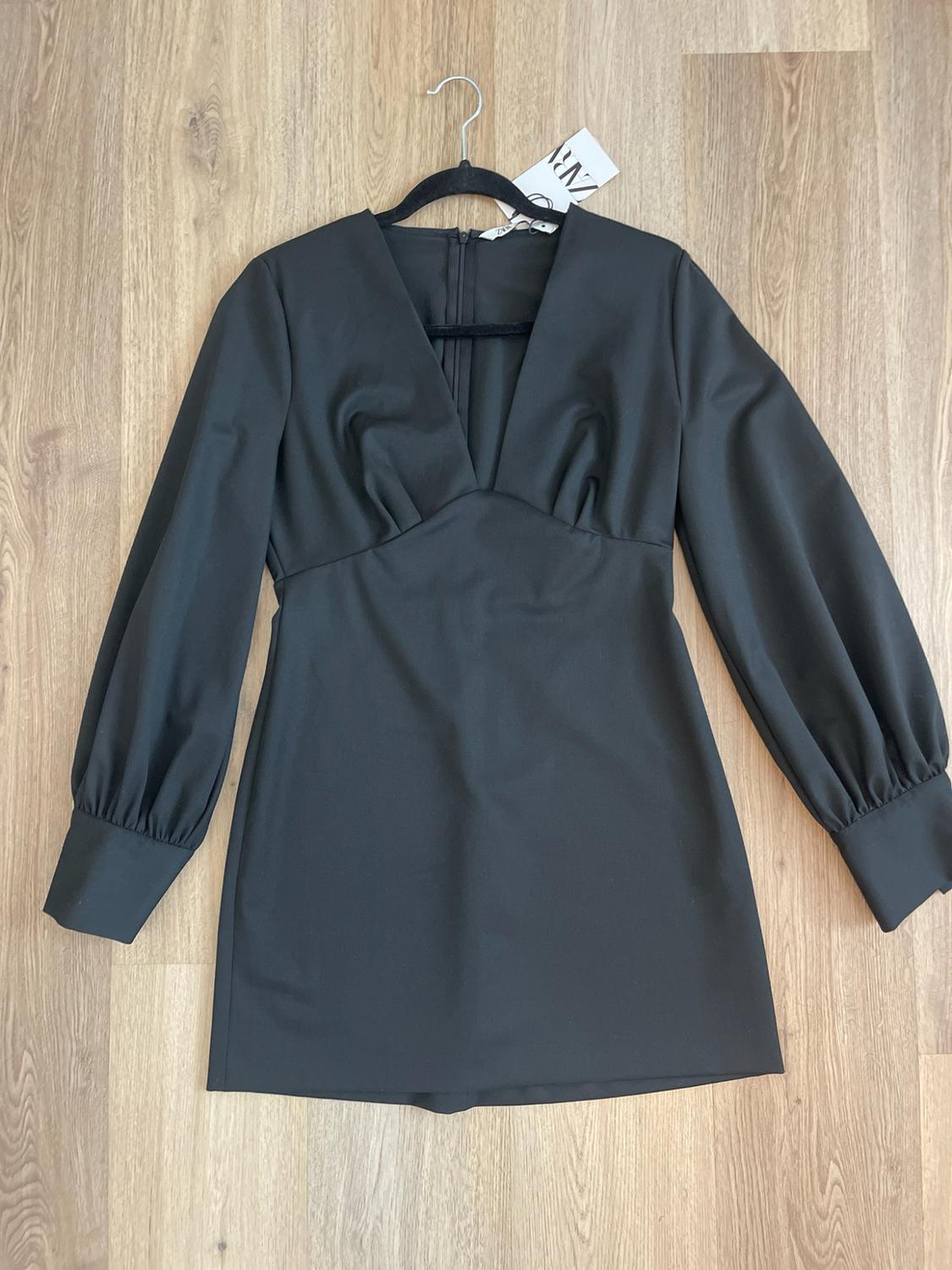 Black short dress with long sleeves and a V-neck, hanging on a hanger against a wood background. A Zara tag is visible.