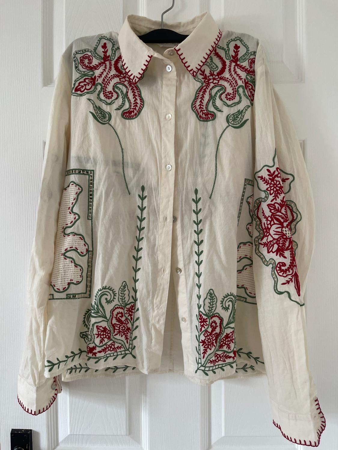 Cream shirt with red and green floral embroidery and red stitching on the collar and cuffs, hanging on a white door.