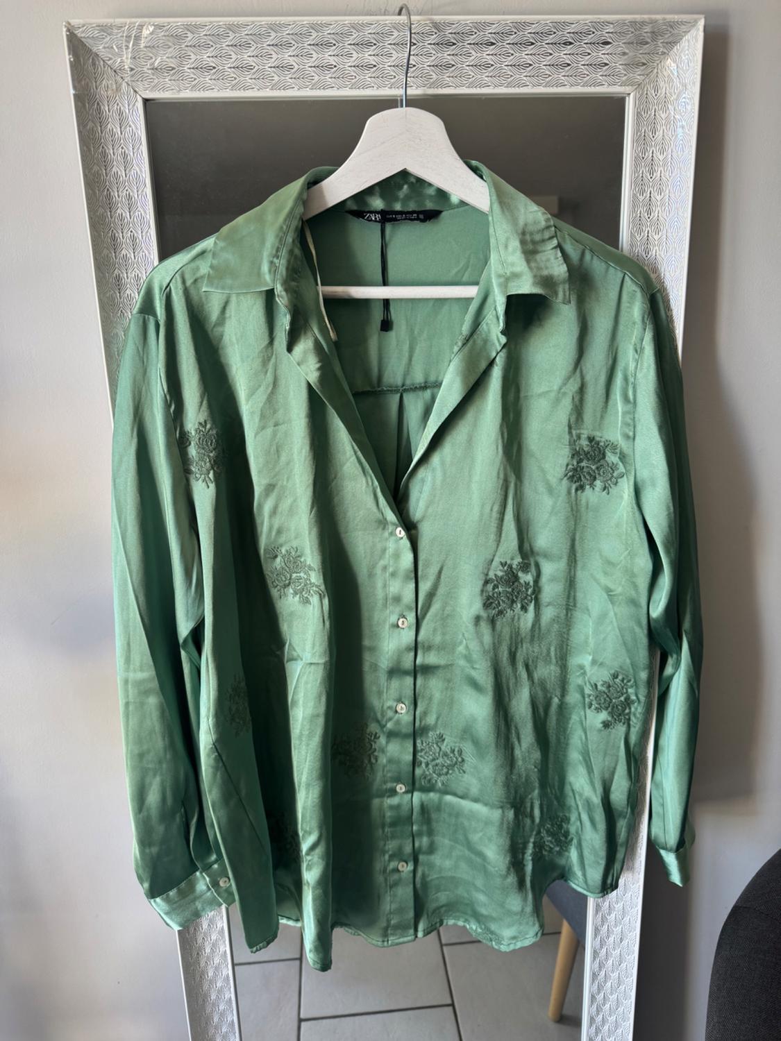Emerald green satin long-sleeve shirt with floral embroidery and a button fastening, hanging on a hanger in front of a mirror.